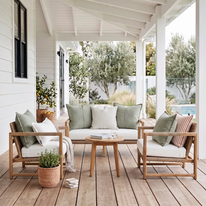 A styled outdoor patio seating set featuring a three-seat sofa, two wooden armchairs, a round wooden coffee table, and multiple decorative throw pillows, arranged on a wooden deck under a white porch with a garden and pool in the background.