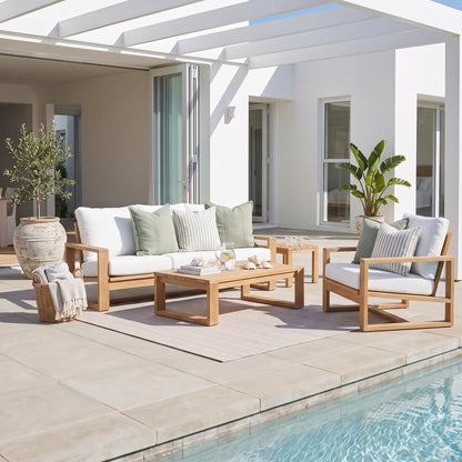 Outdoor patio furniture set including a wooden sofa, matching armchair, and coffee table with white cushions and green pillows, arranged on a light rug beside a swimming pool in a modern white patio setting.
