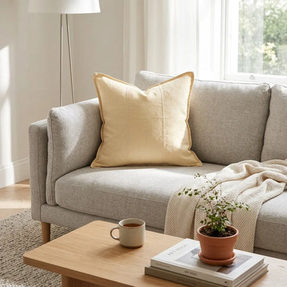 A light beige throw pillow with a subtle stitched pattern and a thin darker border, styled on a light gray fabric sofa in a bright, modern living room setting.