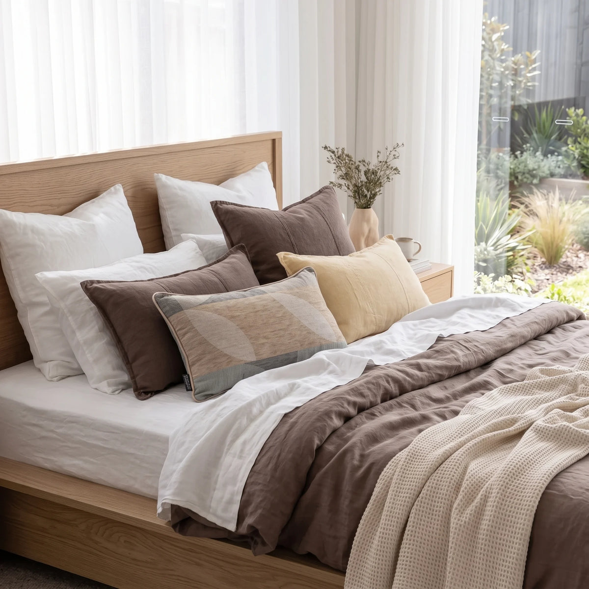 A neatly made bed in a bright bedroom setting, featuring a brown and white bedding set with multiple decorative pillows, a light wood headboard and footboard, sheer white curtains, and a small vase with dried stems on a bedside table.