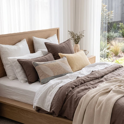 A neatly made bed in a bright bedroom setting, featuring a brown and white bedding set with multiple decorative pillows, a light wood headboard and footboard, sheer white curtains, and a small vase with dried stems on a bedside table.