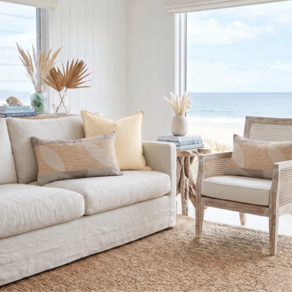A bright coastal living room with a cream sofa, patterned throw pillows, a matching armchair, and a woven rug, with an ocean view visible through large windows.