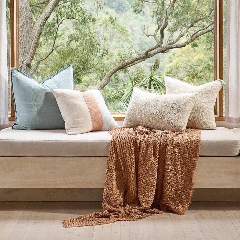 Decorative pillows and a blanket on a window seat with a view of trees.