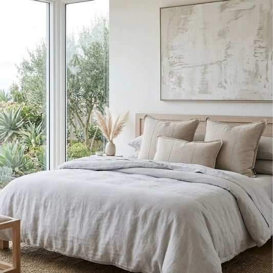 Neatly made bed with beige and gray bedding in a room with large windows and outdoor view.