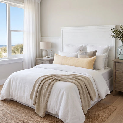 A neatly made bed in a bright, neutral-toned bedroom. The bedding set is primarily white with a textured cream throw blanket and a pale yellow accent pillow.