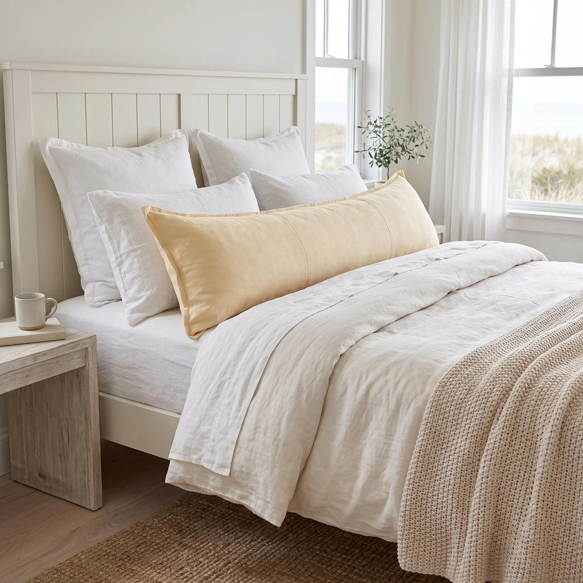 A neatly made bed in a bright bedroom setting, featuring a cream and off-white bedding set with a prominent pale yellow long pillow. The bed has a white slatted headboard, a light wood nightstand with a mug, and a textured beige throw blanket.