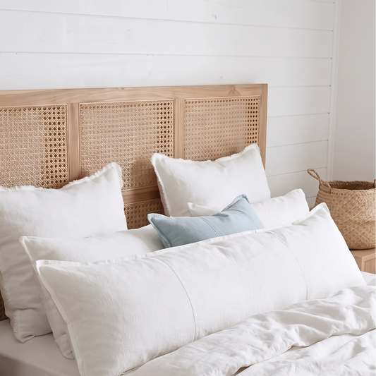 A neatly made bed with white bedding, a light wood headboard with woven cane panels, and a matching wood bedside table holding a lamp and books in a bright bedroom.