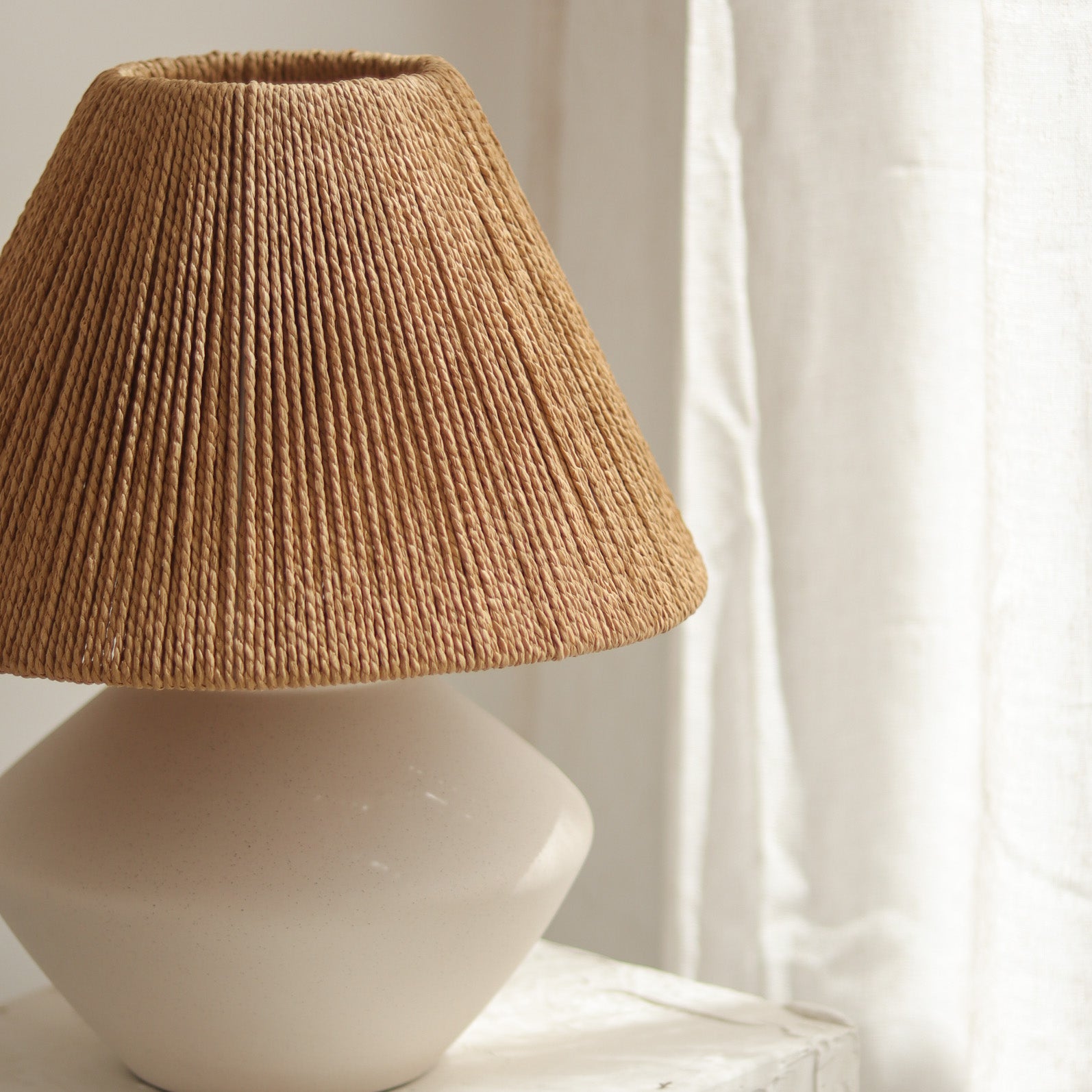 Lamp with a textured raffia shade on a white base against a light curtain background