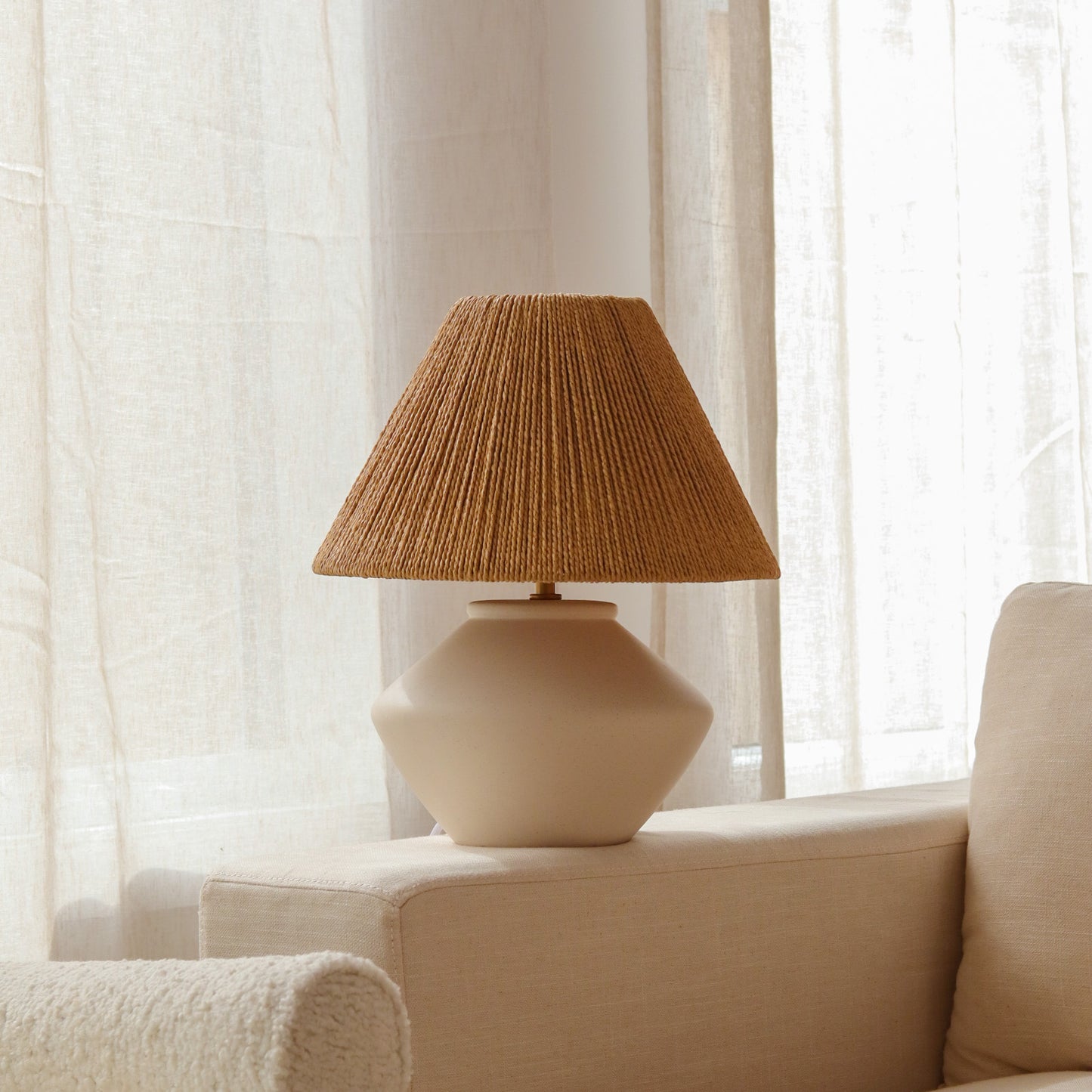 Beige lamp with a raffia shade on a white surface with curtains in the background