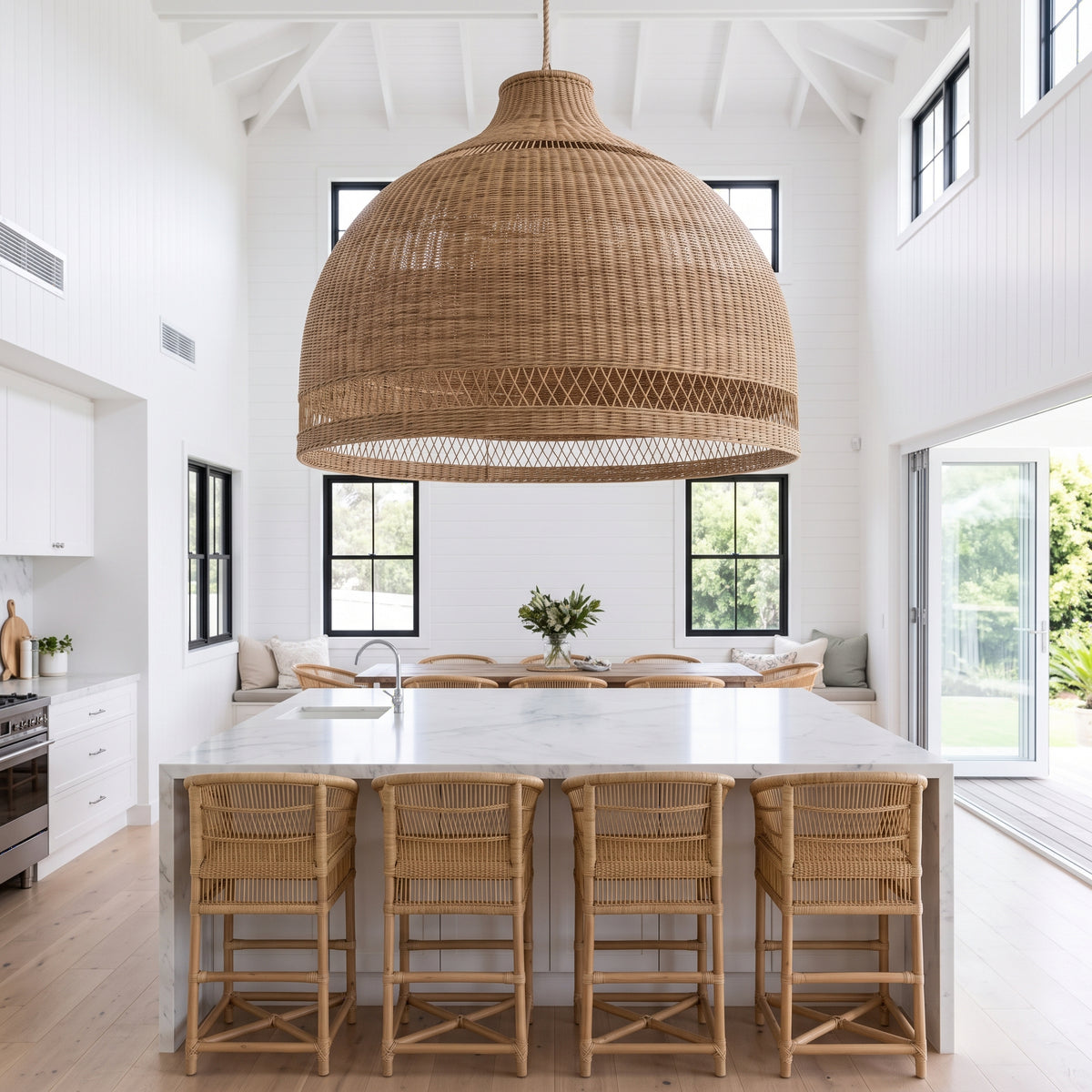 A large woven wicker pendant light in a dome shape hangs over a modern kitchen island with a white marble-look top and four woven bar stools, set in a bright white high-ceilinged kitchen.