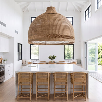 A large woven wicker pendant light in a dome shape hangs over a modern kitchen island with a white marble-look top and four woven bar stools, set in a bright white high-ceilinged kitchen.