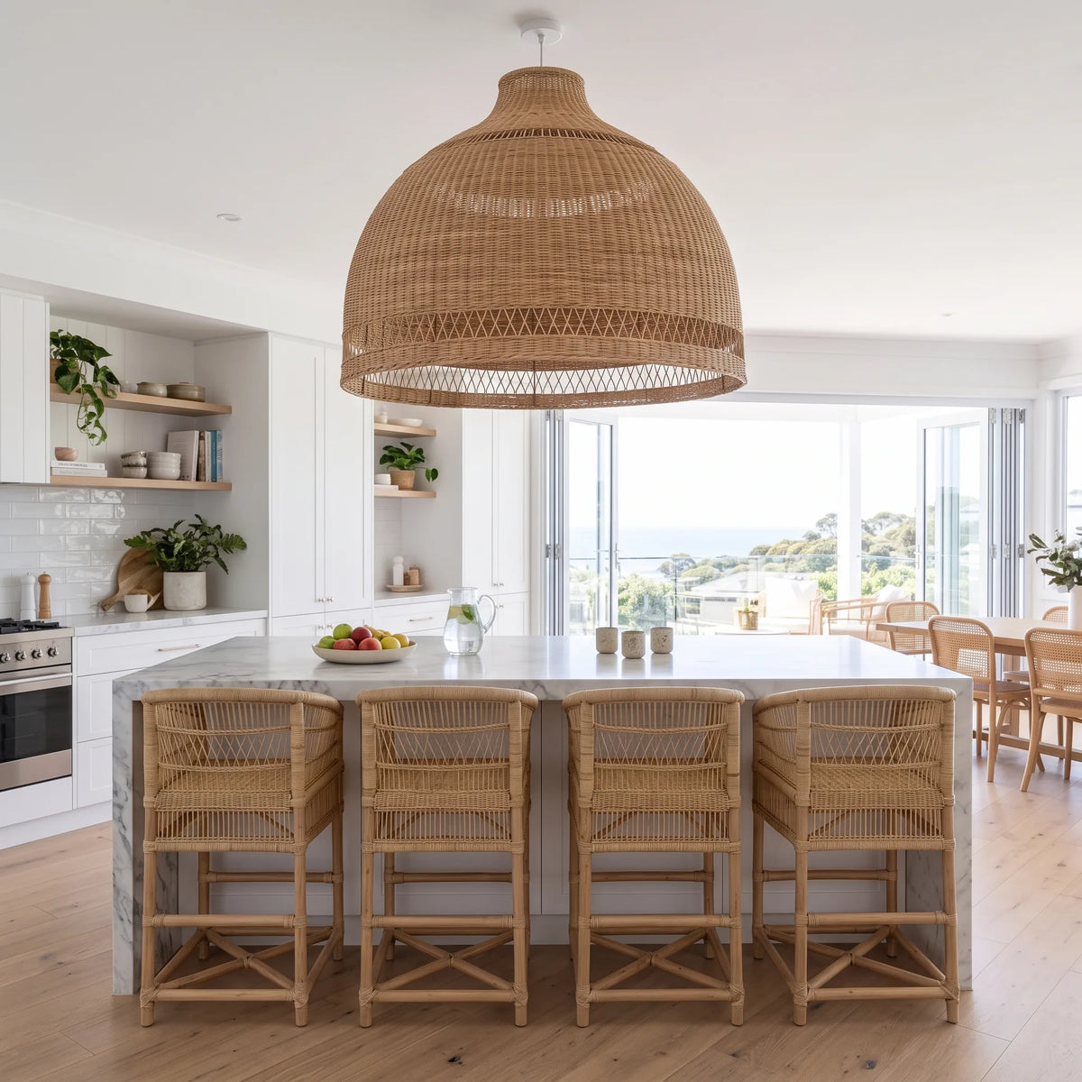 A large woven wicker pendant light hangs over a modern kitchen island with a marble countertop and four matching woven bar stools. The kitchen has white cabinets, open wood shelves with plants, and large windows overlooking a scenic outdoor view.