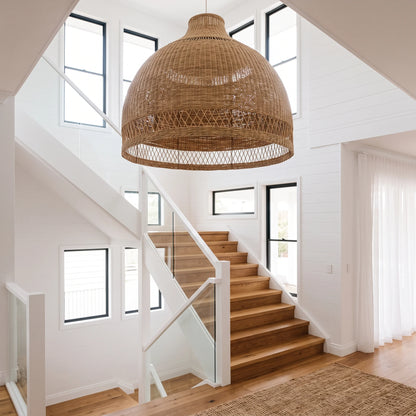 A large woven wicker pendant light in a dome shade hangs over a wooden staircase in a bright, modern home interior with white walls and glass railings.