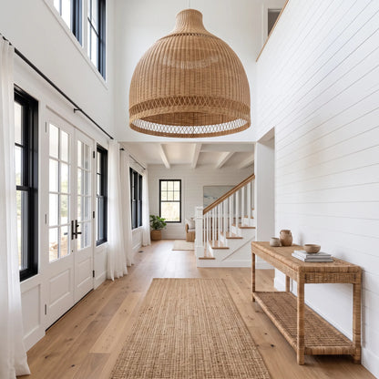 A large woven rattan dome pendant light hangs in a bright, modern white hallway with wood floors, a console table, and a staircase in the background.