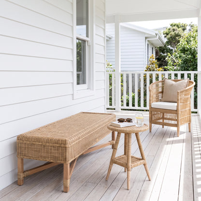 A wicker outdoor bench and matching side table on a white porch, with a woven armchair and a small cushion nearby. The furniture is made of natural tan wicker with light wood legs, set against a white house exterior and light deck flooring.