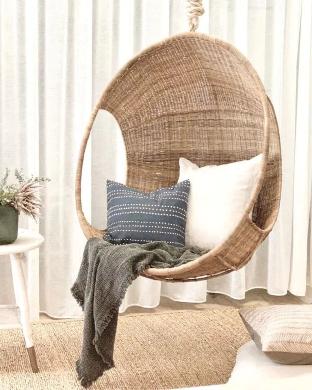 Woven hanging chair with cushions and a blanket in a room with white curtains.