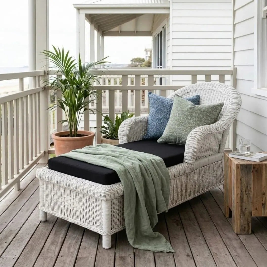 White wicker lounge chair with cushions on a wooden deck, featuring a plant and small table.