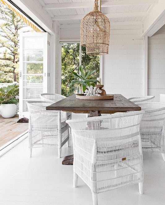Outdoor dining area with wooden table and white wicker chairs, surrounded by greenery.