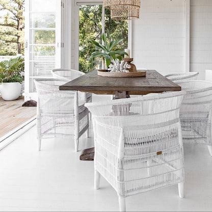 Outdoor dining area with wooden table and white wicker chairs, surrounded by greenery.