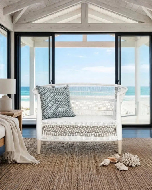 White wicker chair with a blue pillow in a room with large windows overlooking the ocean.