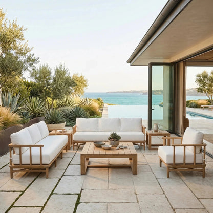 Outdoor patio set with white cushions and teak frames on a stone patio overlooking a pool and ocean view.
