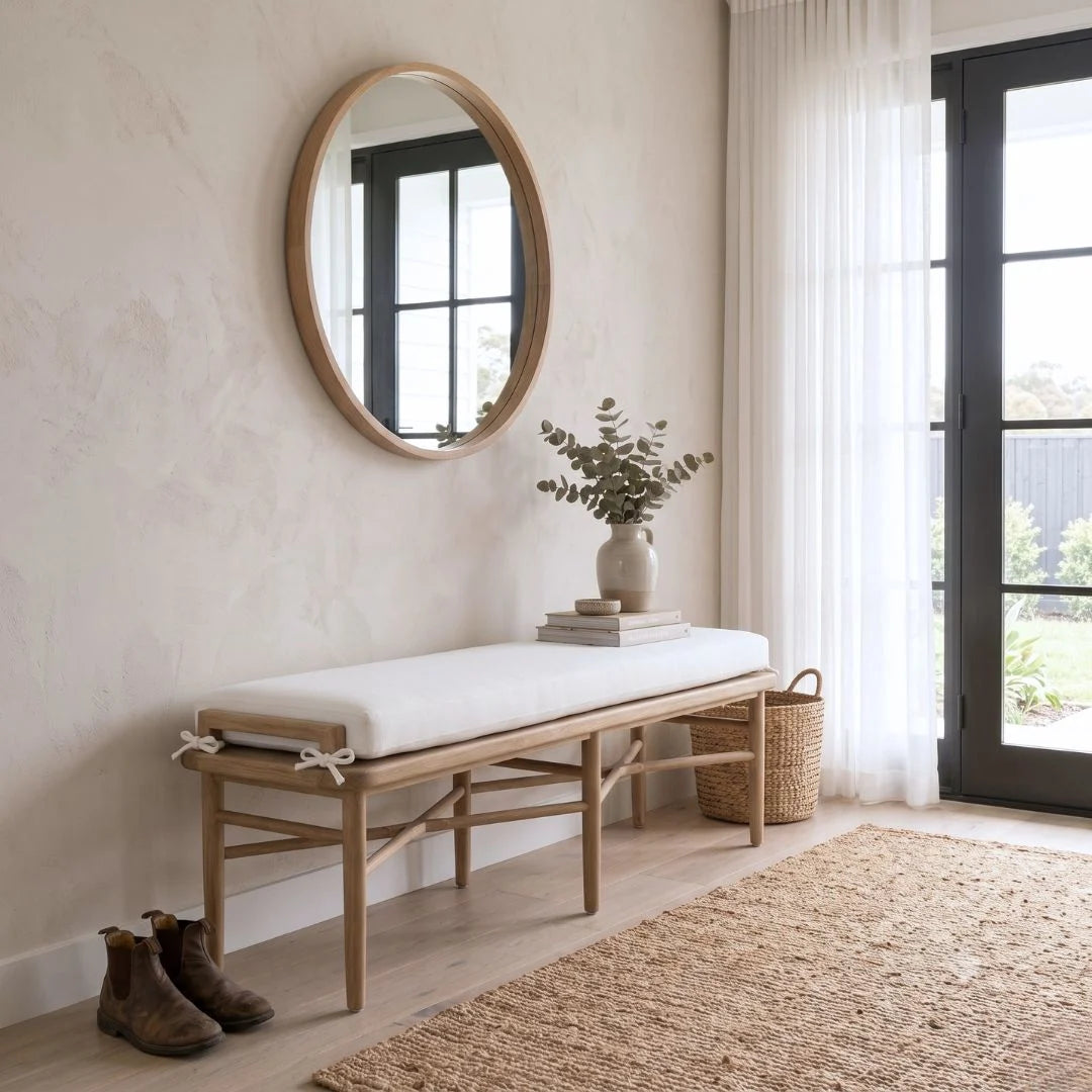 Wooden bench with a white cushion in a room with a round mirror, plant, and door.