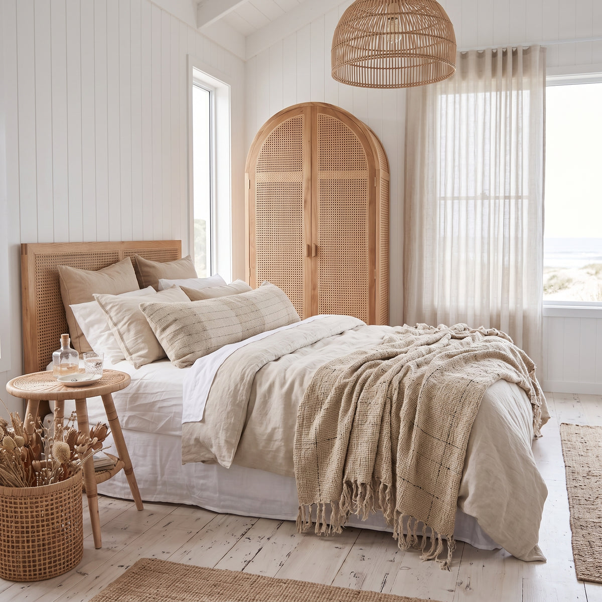 A beautifully styled bohemian bedroom featuring a bed dressed in neutral beige and white linens, a woven rattan wardrobe, a hanging rattan pendant light, and a wicker basket on the floor, all set against white paneled walls.