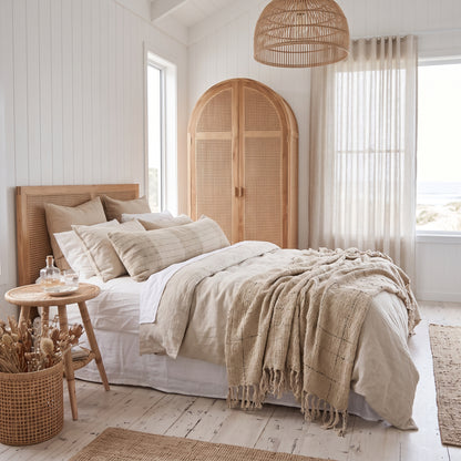 A beautifully styled bohemian bedroom featuring a bed dressed in neutral beige and white linens, a woven rattan wardrobe, a hanging rattan pendant light, and a wicker basket on the floor, all set against white paneled walls.