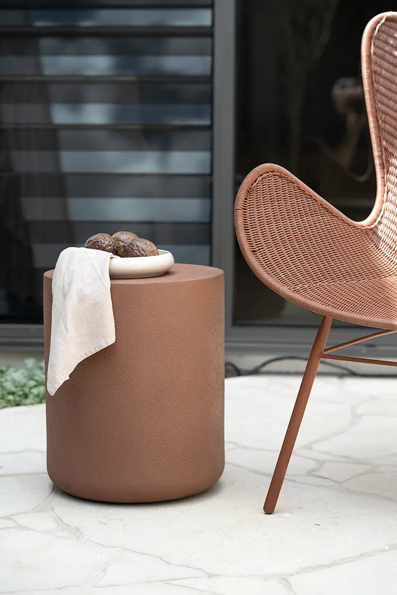 Brown cylindrical side table with a white cloth and decorative items next to a wicker chair outdoors.