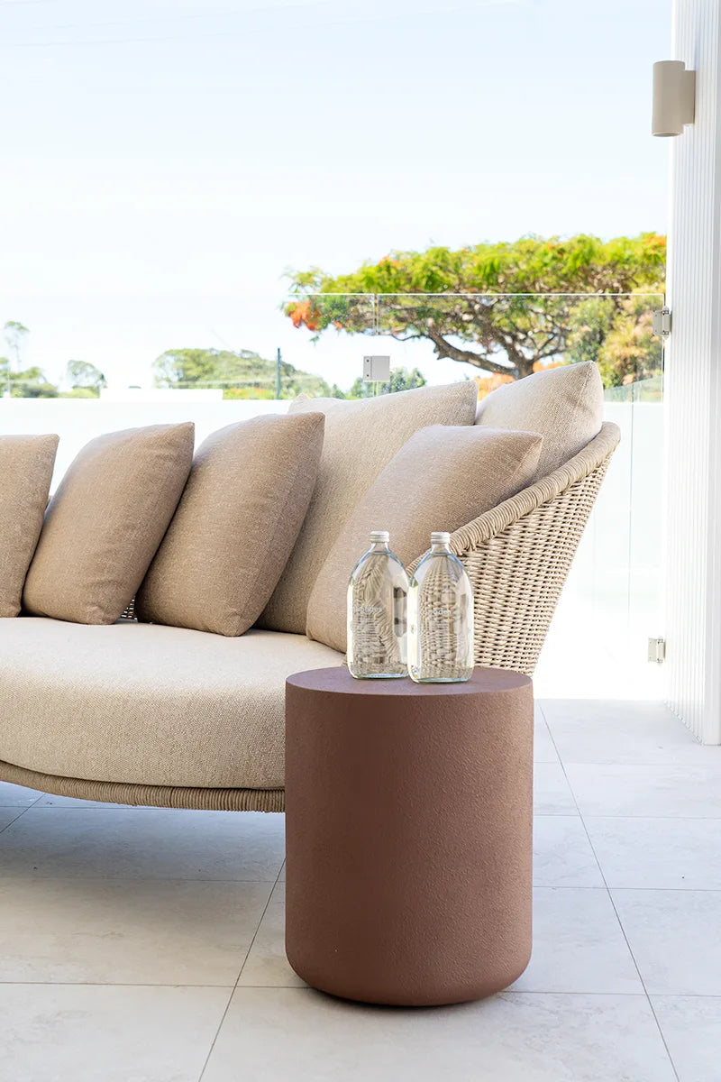 Beige outdoor sofa with a woven backrest and a small round brown table with decorative bottles on a patio.