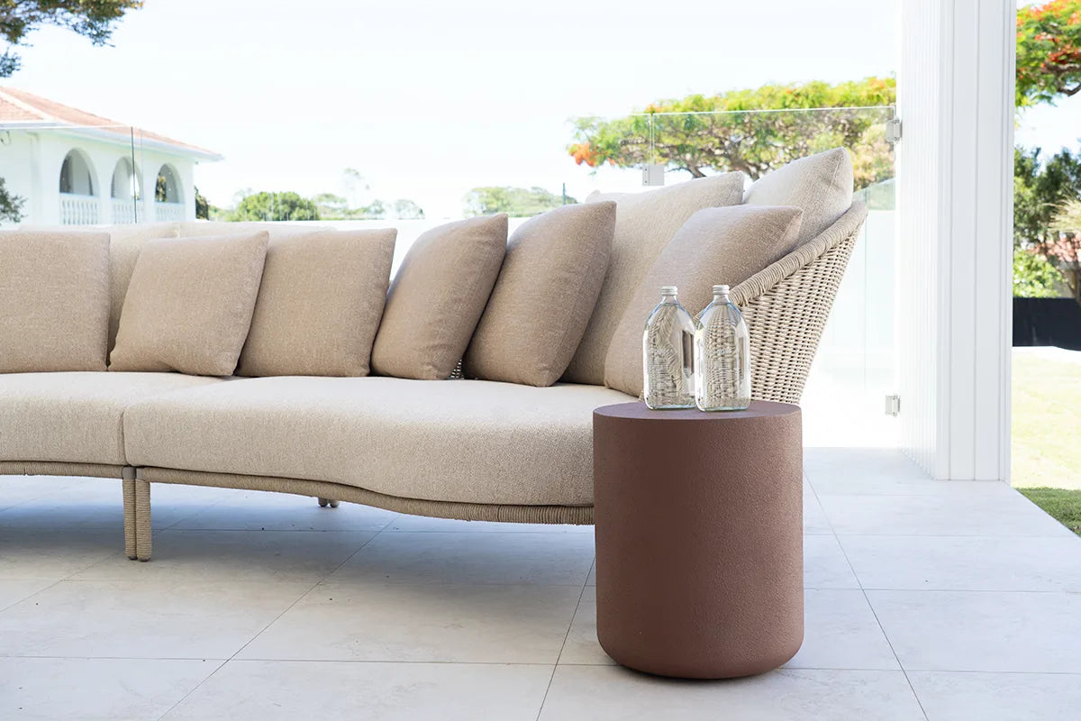 Beige outdoor sofa with a cylindrical side table and two bottles on a patio.
