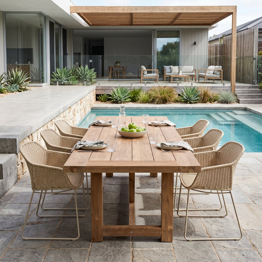 A rectangular wooden dining table with six woven wicker chairs is set on a stone patio next to a swimming pool in a modern outdoor patio setting.