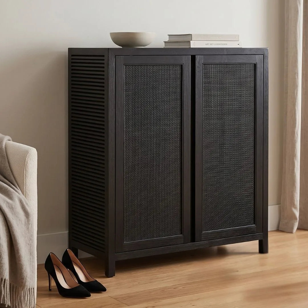 A dark charcoal gray wooden cabinet with woven cane doors, styled in a minimalist room with a bowl and books on top and black heels on the floor.