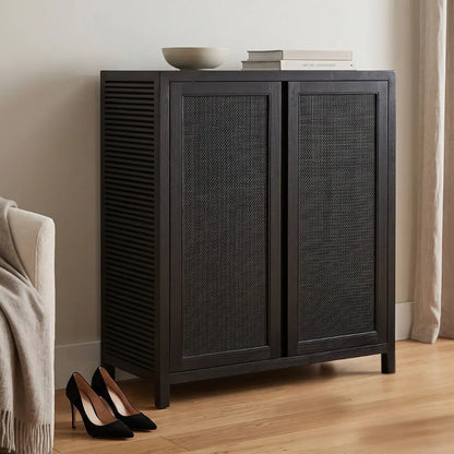 A dark charcoal gray wooden cabinet with woven cane doors, styled in a minimalist room with a bowl and books on top and black heels on the floor.