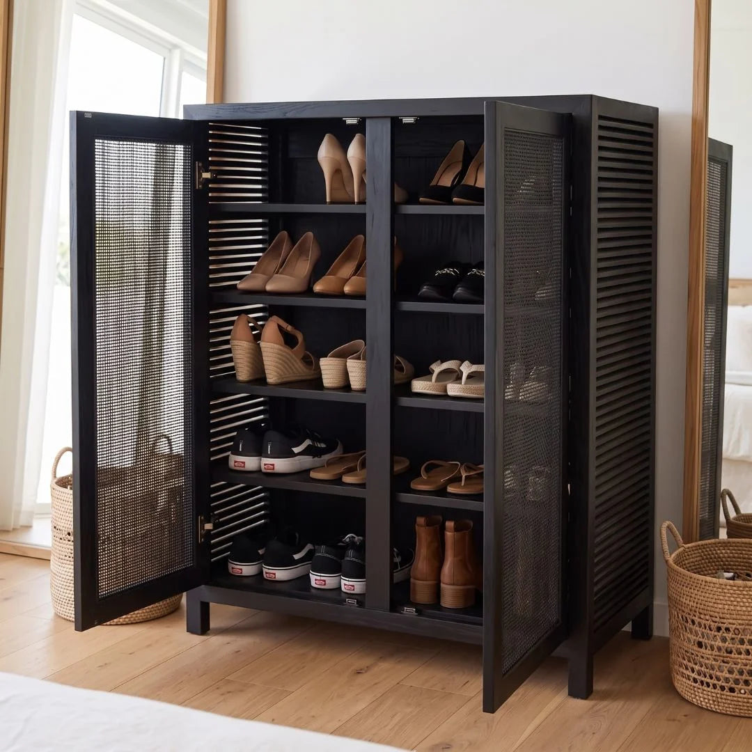 A tall black shoe cabinet with two open doors, showing multiple shelves filled with shoes and sandals. The cabinet has mesh side panels and is placed in a bright room with a wood floor, window, and woven baskets nearby.