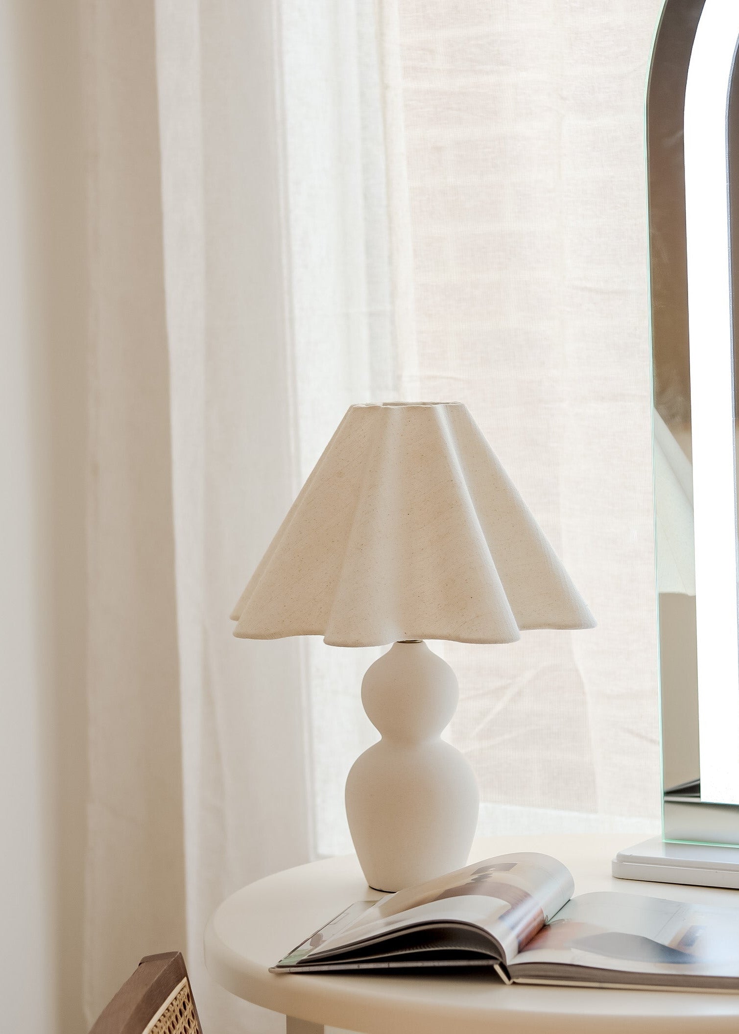 White table lamp on a side table with a magazine against a window with sheer curtains