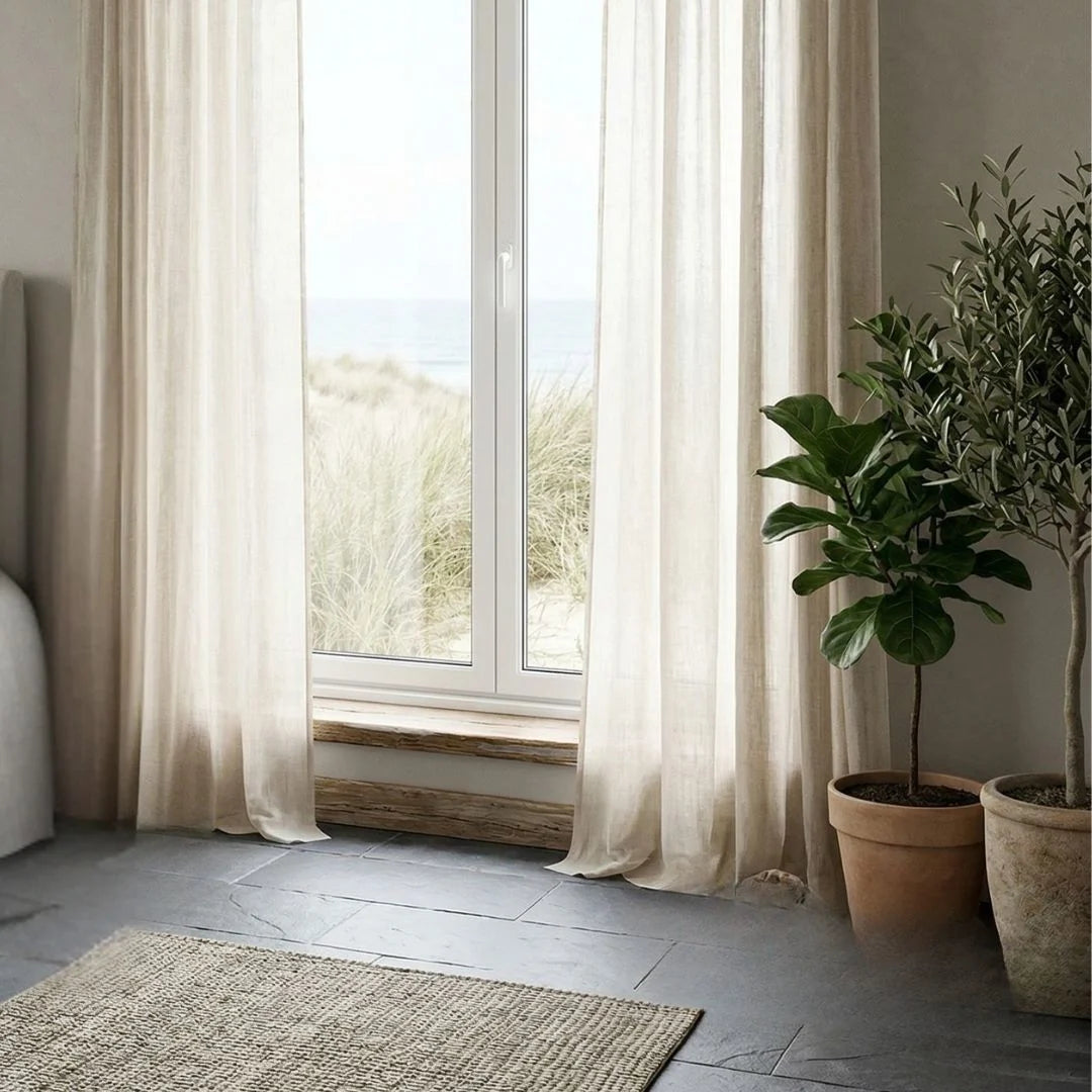 Living room with large windows, white curtains, and various potted plants.