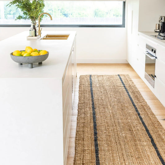 Kitchen with a woven runner rug on a wooden floor, white countertops, and a window in the background.