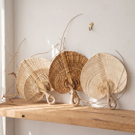 Three woven straw wall fans in natural beige and tan tones displayed on a rustic wooden shelf against a white wall.