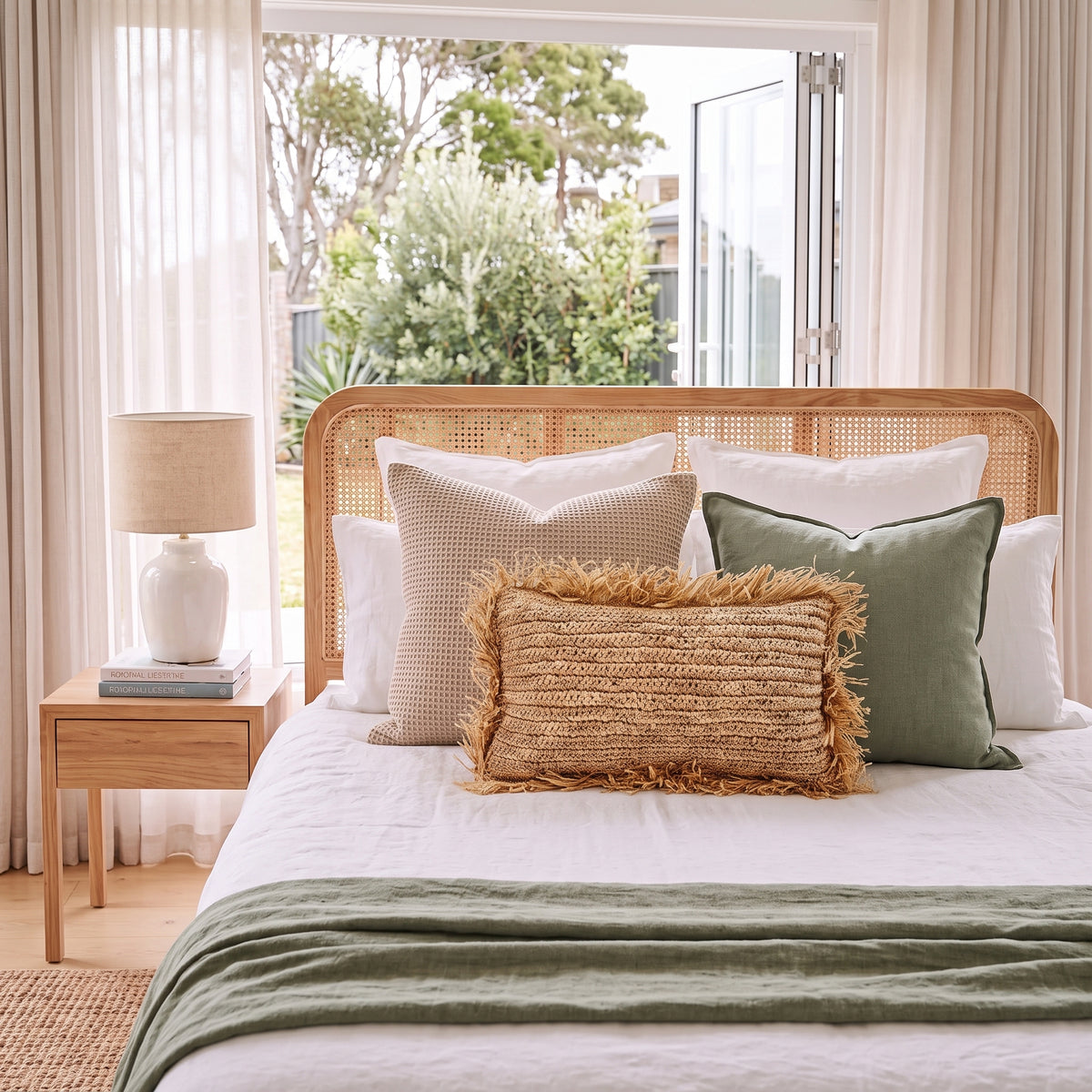 A styled bed in a bright bedroom with a rattan headboard, white bedding, and three decorative pillows in beige, green, and textured tan. A matching green throw blanket is draped at the foot of the bed, with a wooden side table and lamp on the left and a large window behind showing greenery outside.