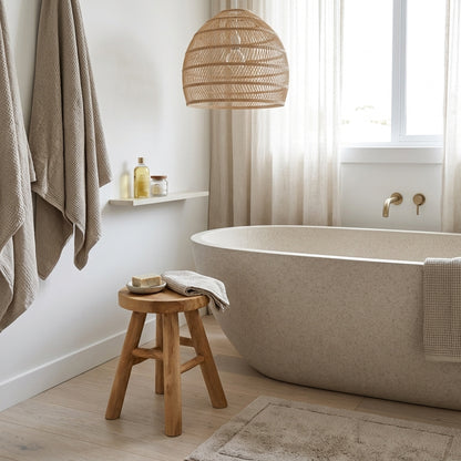 A modern freestanding bathtub in a light beige stone finish, styled in a bright, minimalist bathroom with a woven pendant light, wooden stool, and hanging towels.