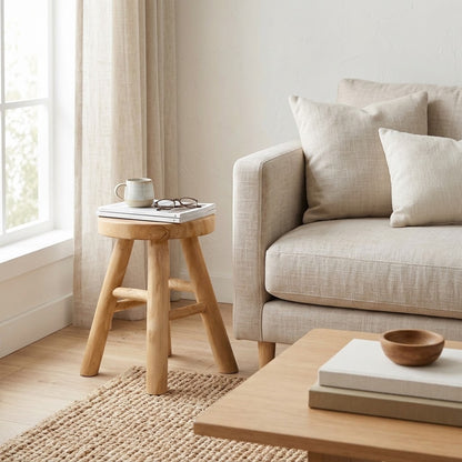 A light natural wood three-legged stool with a small footrest, styled in a bright living room next to a beige sofa.