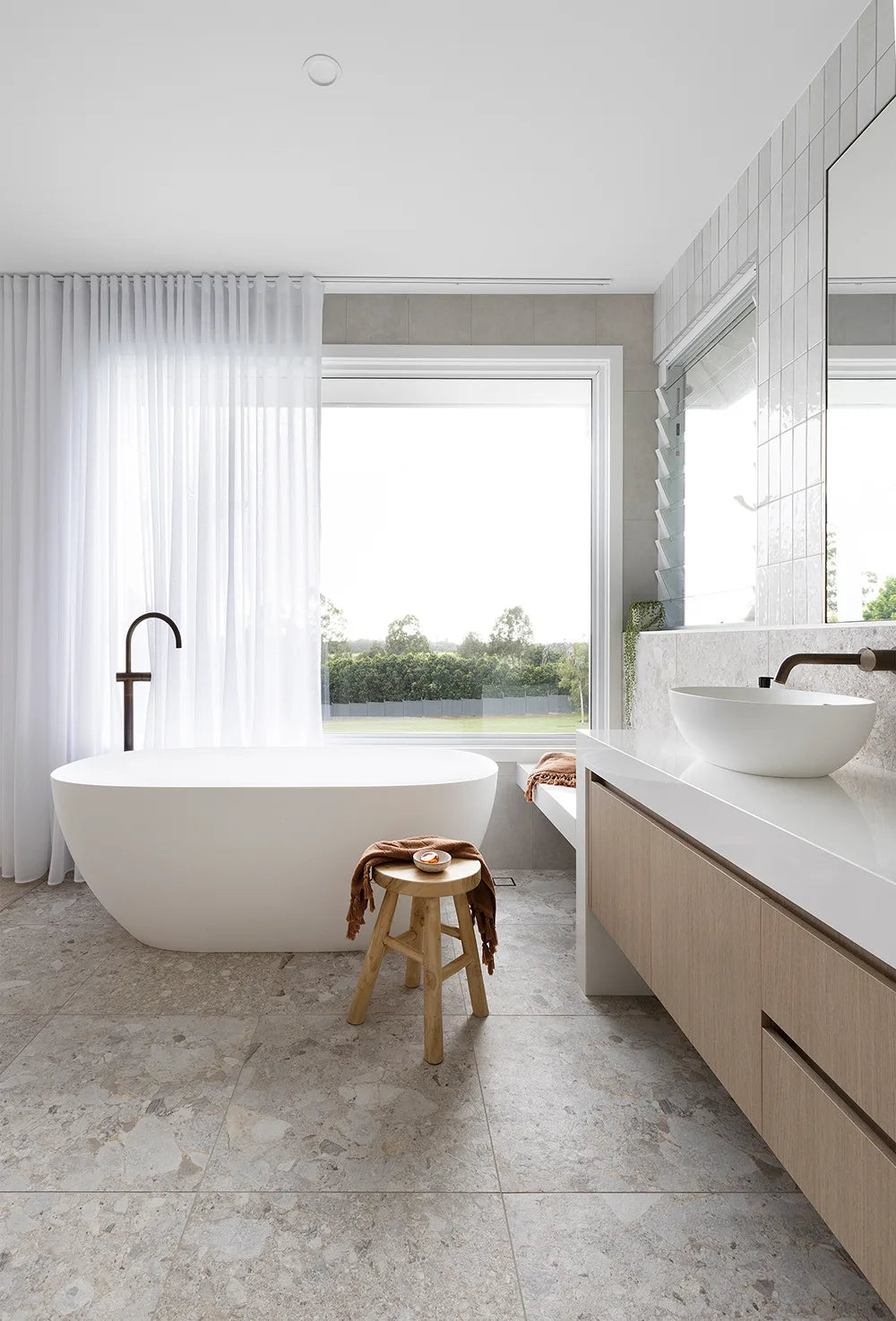 Modern bathroom with a freestanding bathtub, wooden stool, and large window.