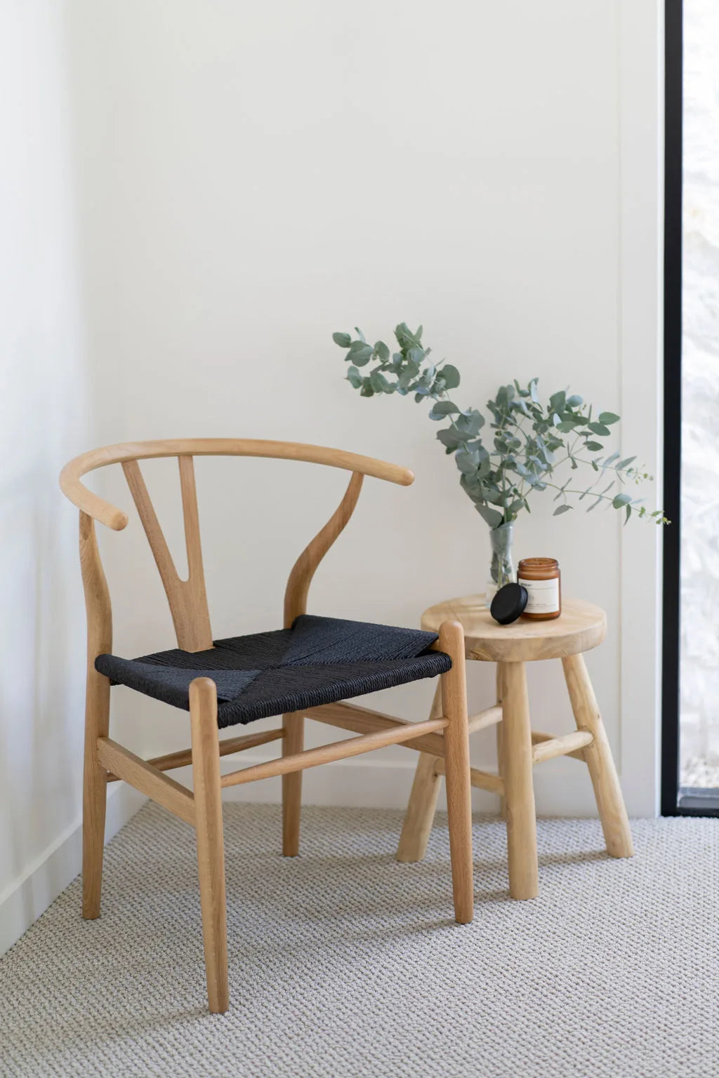 Wishbone chair with black seat and a small teak table with a plant and candle in a minimalistic room.