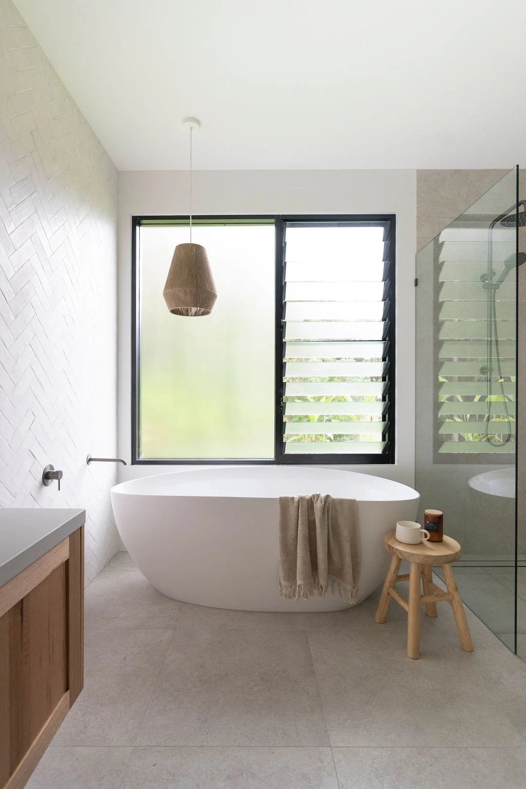Modern bathroom with a freestanding bathtub, wooden stool, and window with blinds.