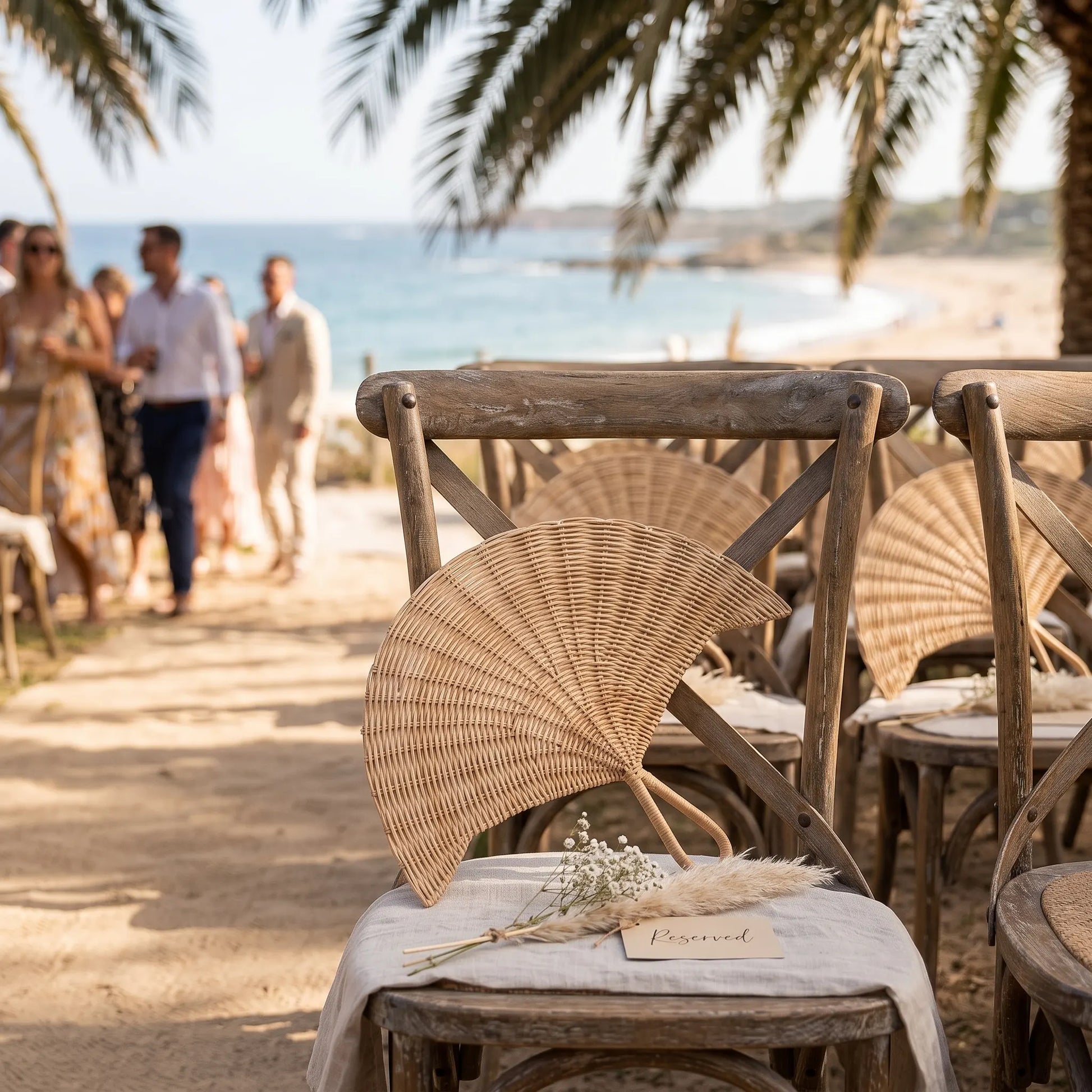 Beach-side wooden chairs with woven rattan fans and draped white fabric, set in a sandy outdoor venue with palm trees and guests walking in the background near the ocean.