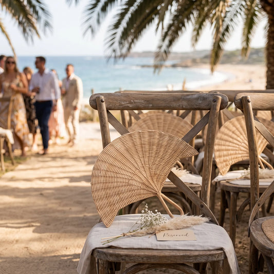 Beach-side wooden chairs with woven rattan fans and draped white fabric, set in a sandy outdoor venue with palm trees and guests walking in the background near the ocean.