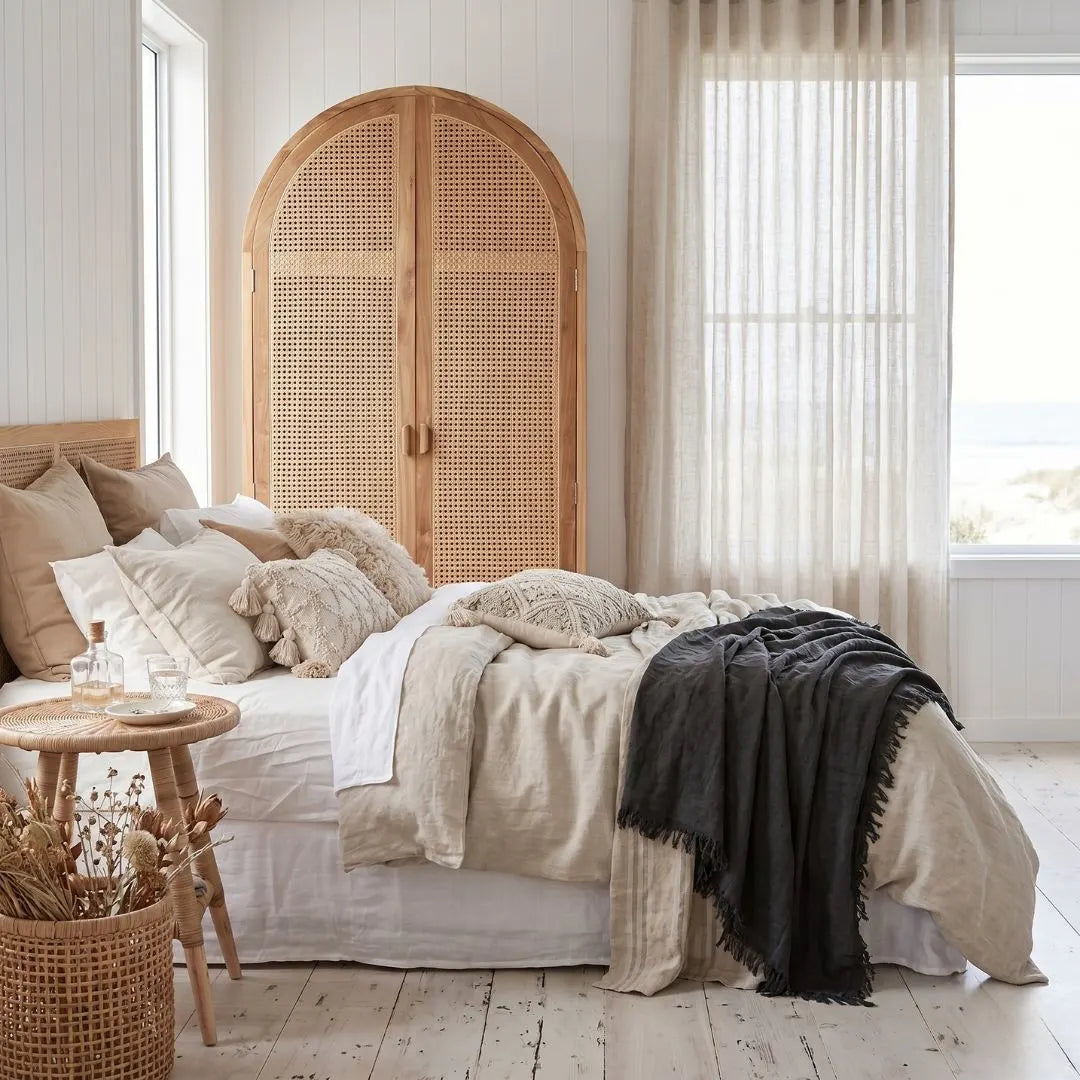 Neatly made bed with beige bedding and a black throw blanket in a bedroom with wooden wardrobe and large windows.