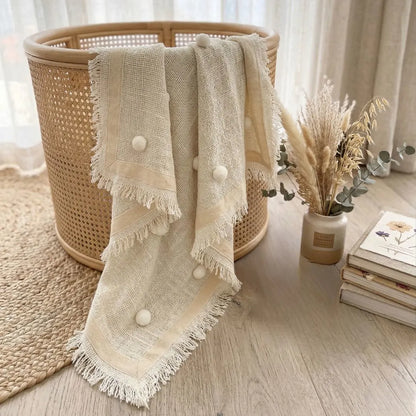 Beige textured blanket with white pom-poms draped over a woven basket on a wooden floor.