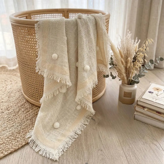 Beige textured blanket with white pom-poms draped over a woven basket on a wooden floor.